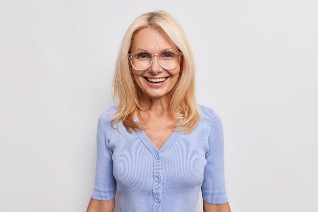 woman-with-blonde-hair-smiles-joyfully-enjoys-life-wears-big-optical-glasses-blue-jumper-happy-guests-came-isolated-white-people-age_273609-56429